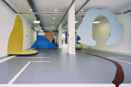 An art installation by Eva Funk with large abstract shapes in red, yellow, blue, and purple, displayed in an industrial-style gallery space. White walls and geometric patterns on the floor. Artist  Eva Funk