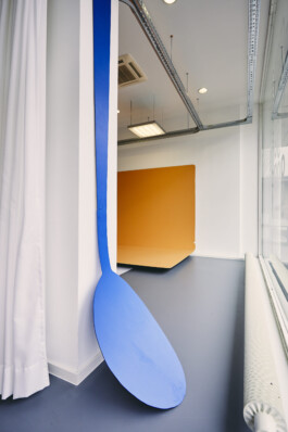 An art installation by Eva Funk with large abstract shapes in red, yellow, blue, and purple, displayed in an industrial-style gallery space. White walls and geometric patterns on the floor. Artist  Eva Funk