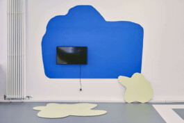 An art installation by Eva Funk with large abstract shapes in red, yellow, blue, and purple, displayed in an industrial-style gallery space. White walls and geometric patterns on the floor. Artist  Eva Funk