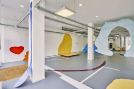 An art installation by Eva Funk with large abstract shapes in red, yellow, blue, and purple, displayed in an industrial-style gallery space. White walls and geometric patterns on the floor. Artist  Eva Funk