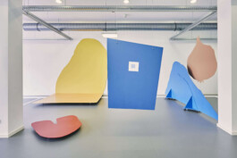 An art installation by Eva Funk with large abstract shapes in red, yellow, blue, and purple, displayed in an industrial-style gallery space. White walls and geometric patterns on the floor. Artist  Eva Funk
